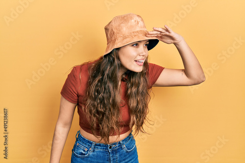 Young hispanic girl wearing casual clothes and hat very happy and smiling looking far away with hand over head. searching concept.