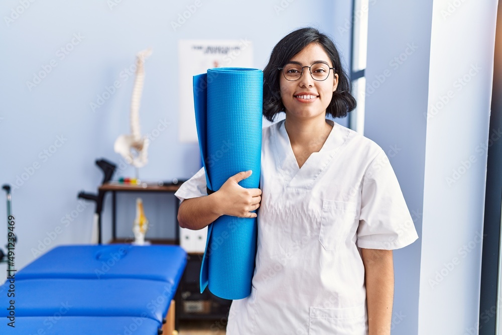Young latin woman wearing physiotherapist uniform holding yoga mat at physiotherapy clinic