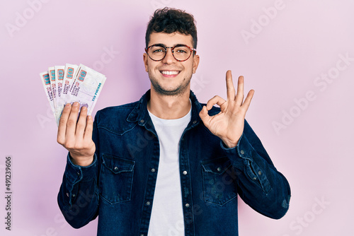 Young hispanic man holding egyptian pounds banknotes doing ok sign with fingers, smiling friendly gesturing excellent symbol photo
