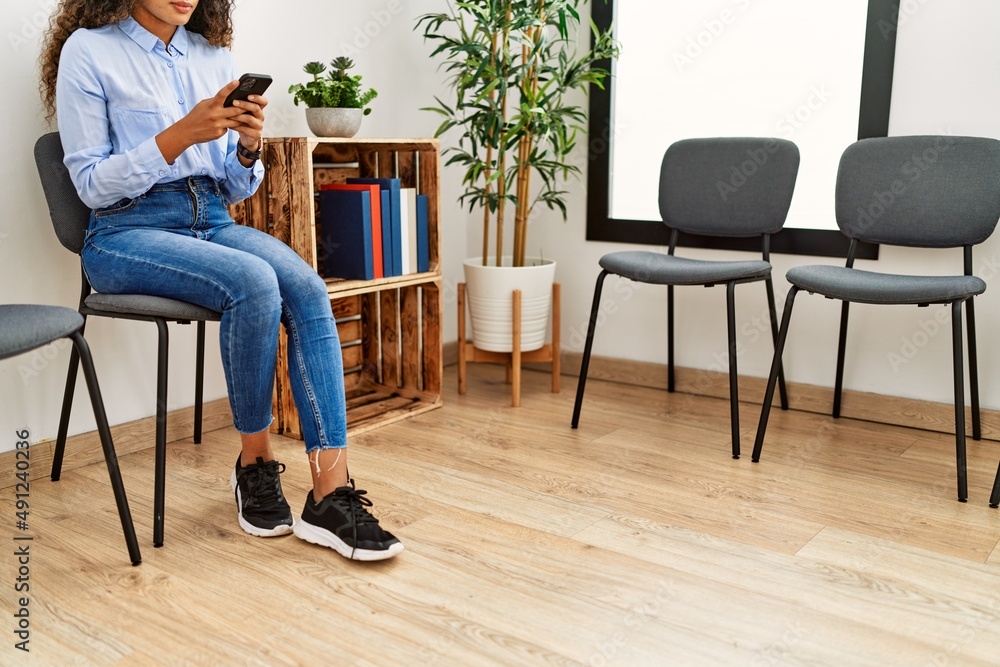 Young latin woman using smartphone sitting on chair at waiting room