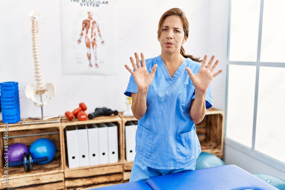 Middle age hispanic physiotherapist woman working at pain recovery clinic moving away hands palms showing refusal and denial with afraid and disgusting expression. stop and forbidden. Middle age hispanic physiotherapist woman working at pain recovery clinic moving away hands palms showing refusal and denial with afraid and disgusting expression. stop and forbidden.