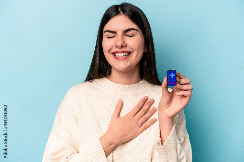 Young caucasian woman holding a batterie isolated on blue background laughs out loudly keeping hand on chest. Young caucasian woman holding a batterie isolated on blue background laughs out loudly keeping hand on chest.