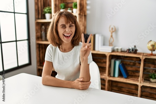 Young caucasian girl wearing casual clothes sitting on the table at home smiling with happy face winking at the camera doing victory sign. number two.