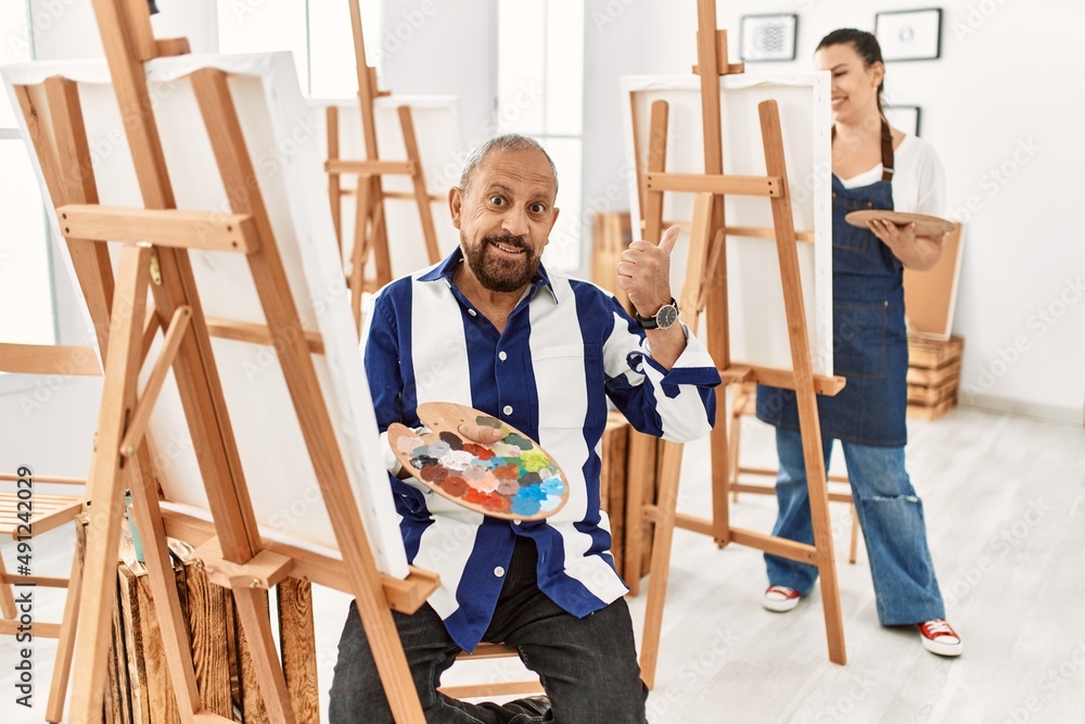 Senior artist man at art studio doing happy thumbs up gesture with hand. approving expression looking at the camera showing success. Senior artist man at art studio doing happy thumbs up gesture with hand. approving expression looking at the camera showing success.