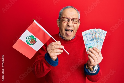 Handsome senior man with grey hair holding peru flag and peruvian sol banknotes sticking tongue out happy with funny expression.
