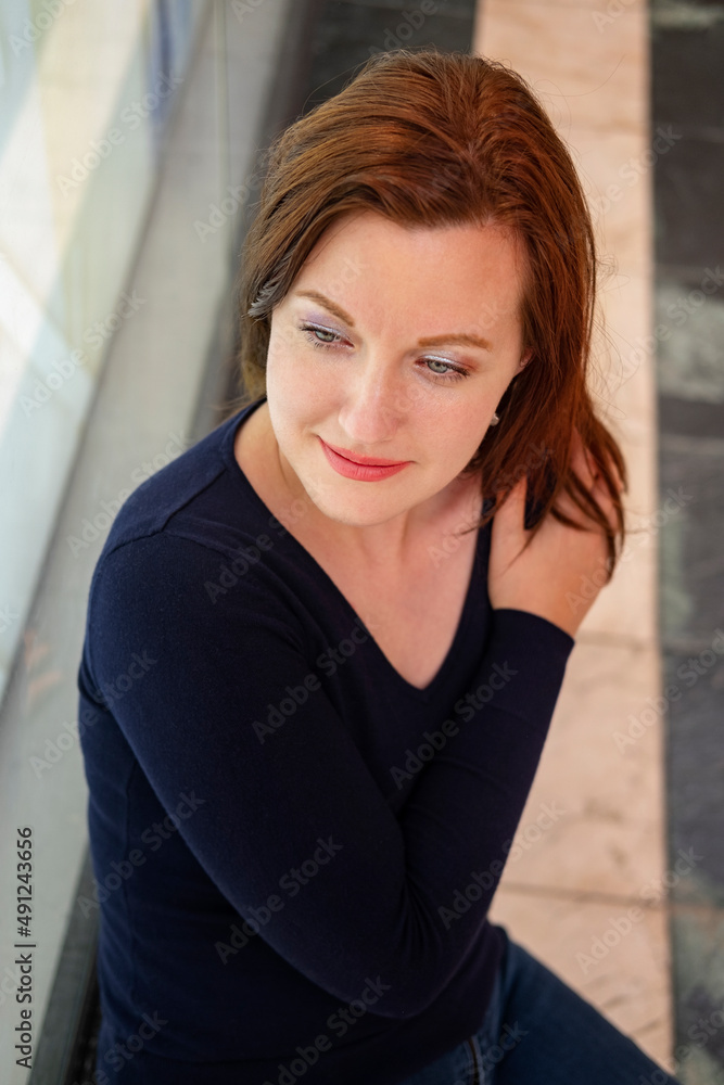 Beautiful Caucasian millennial woman wearing a dark blue velvety sweater and holding her hand and arm across her chest, while sitting on a stone bench and glancing contemplatively with a fading smile Beautiful Caucasian millennial woman wearing a dark blue velvety sweater and holding her hand and arm across her chest, while sitting on a stone bench and glancing contemplatively with a fading smile