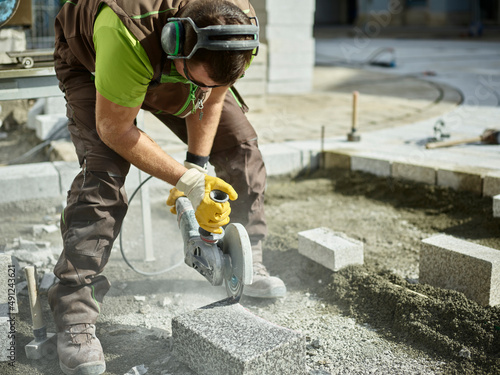 Blue-collar worker cutting paving stone by electric saw at construction site photo