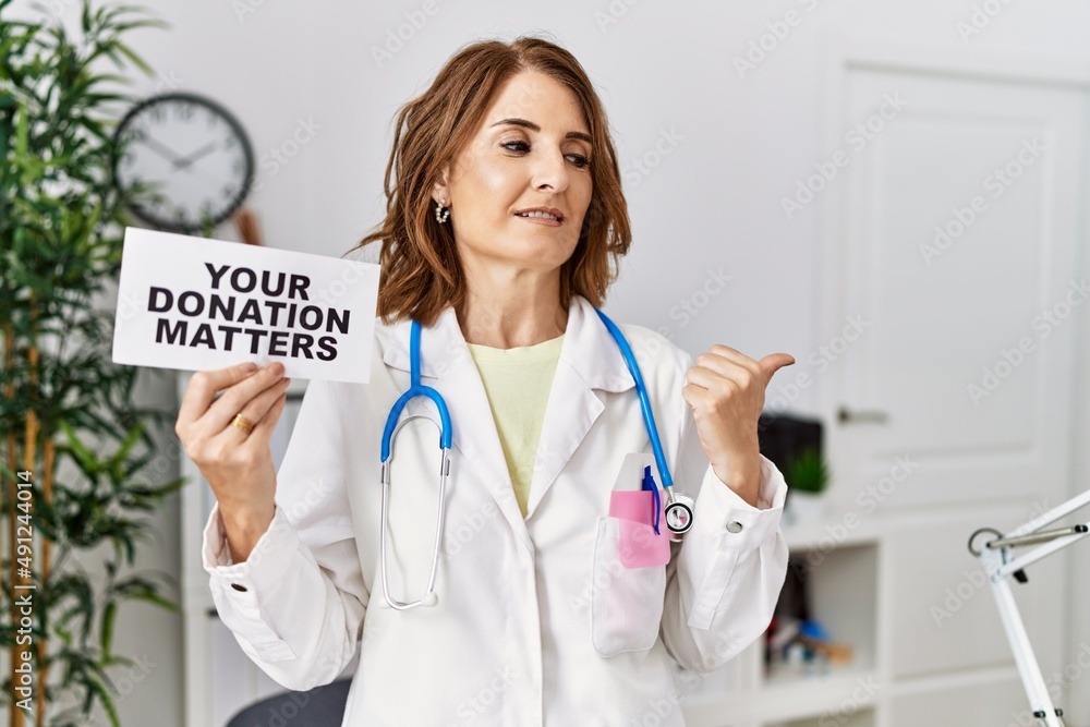 Middle age doctor woman holding your donation matters at the clinic pointing thumb up to the side smiling happy with open mouth Middle age doctor woman holding your donation matters at the clinic pointing thumb up to the side smiling happy with open mouth