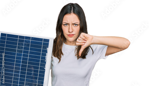 Young beautiful girl holding photovoltaic solar panel with angry face, negative sign showing dislike with thumbs down, rejection concept