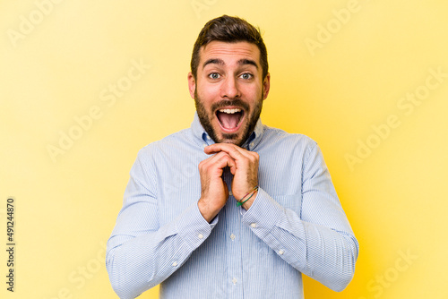 Young caucasian man isolated on yellow background praying for luck, amazed and opening mouth looking to front.