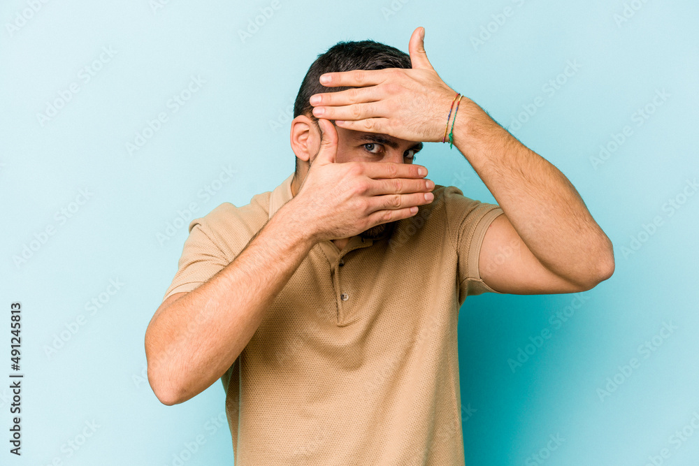 Fototapeta premium Young caucasian man isolated on blue background blink at the camera through fingers, embarrassed covering face.