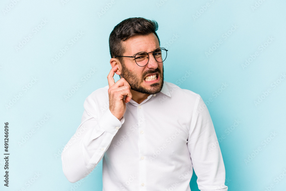 Young caucasian man isolated on blue background covering ears with fingers, stressed and desperate by a loudly ambient. Young caucasian man isolated on blue background covering ears with fingers, stressed and desperate by a loudly ambient.