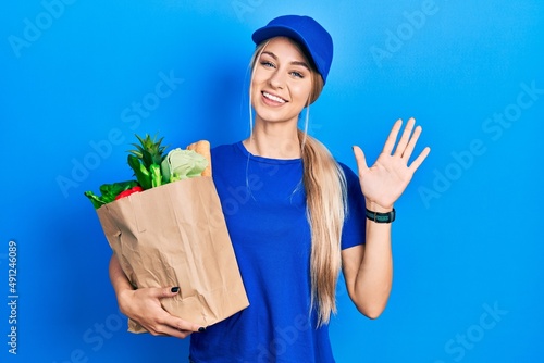 Young caucasian woman wearing courier uniform with groceries from supermarket waiving saying hello happy and smiling, friendly welcome gesture
