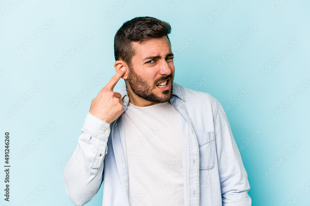 Young caucasian man isolated on blue background covering ears with hands. Young caucasian man isolated on blue background covering ears with hands.