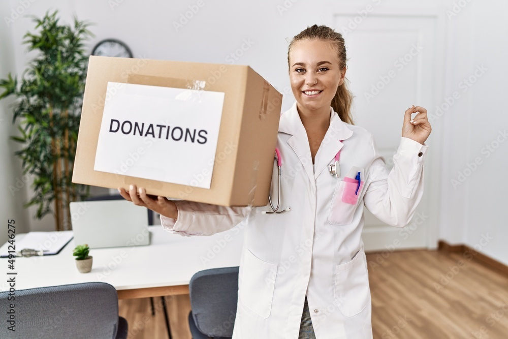 Young doctor woman at the clinic holding donations box smiling happy pointing with hand and finger to the side Young doctor woman at the clinic holding donations box smiling happy pointing with hand and finger to the side