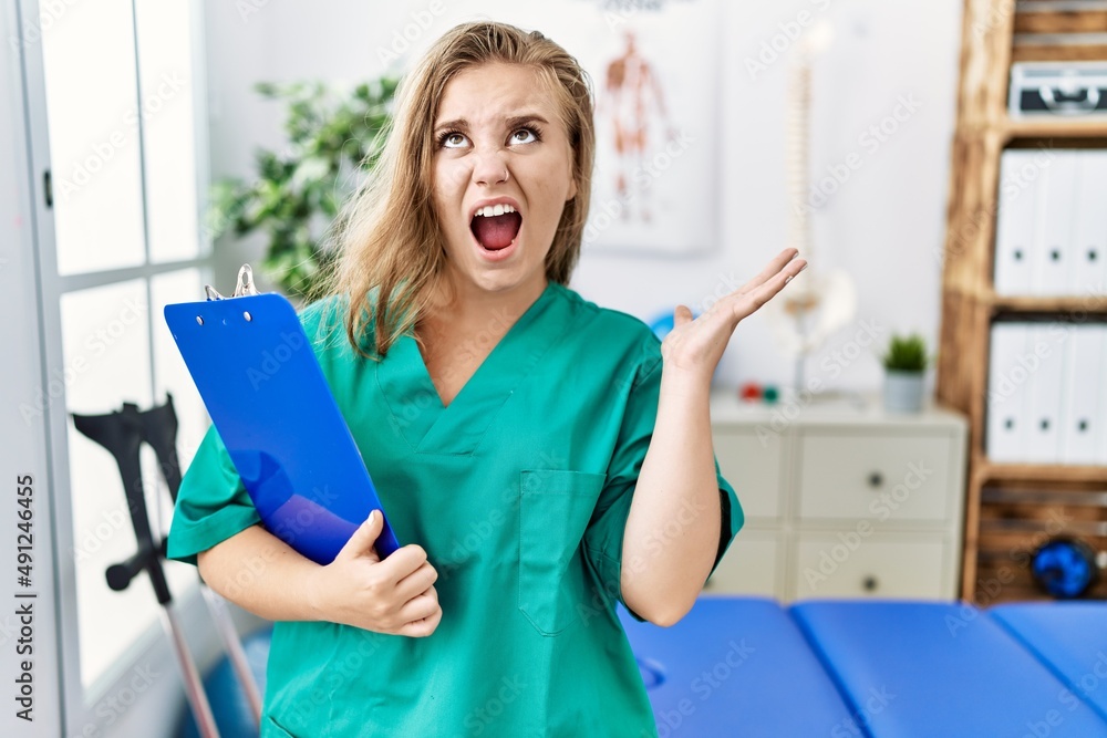 Young caucasian woman working at pain recovery clinic crazy and mad shouting and yelling with aggressive expression and arms raised. frustration concept. Young caucasian woman working at pain recovery clinic crazy and mad shouting and yelling with aggressive expression and arms raised. frustration concept.