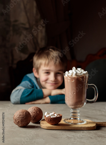 A blond boy with a smile looks at a glass of cocoa or hot chocolate with marshmallows. Chocolate bombs for making cocoa. Selective focus