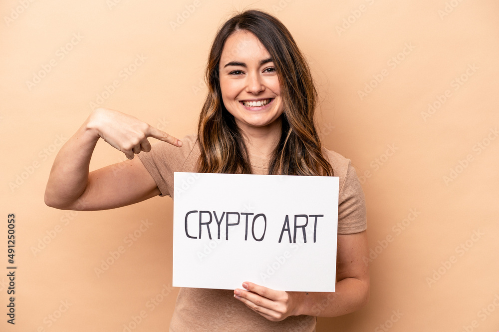 Young caucasian woman holding a crypto art placard isolated on beige background person pointing by hand to a shirt copy space, proud and confident Young caucasian woman holding a crypto art placard isolated on beige background person pointing by hand to a shirt copy space, proud and confident