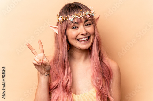 Young elf woman with pink hair isolated on beige background joyful and carefree showing a peace symbol with fingers.
