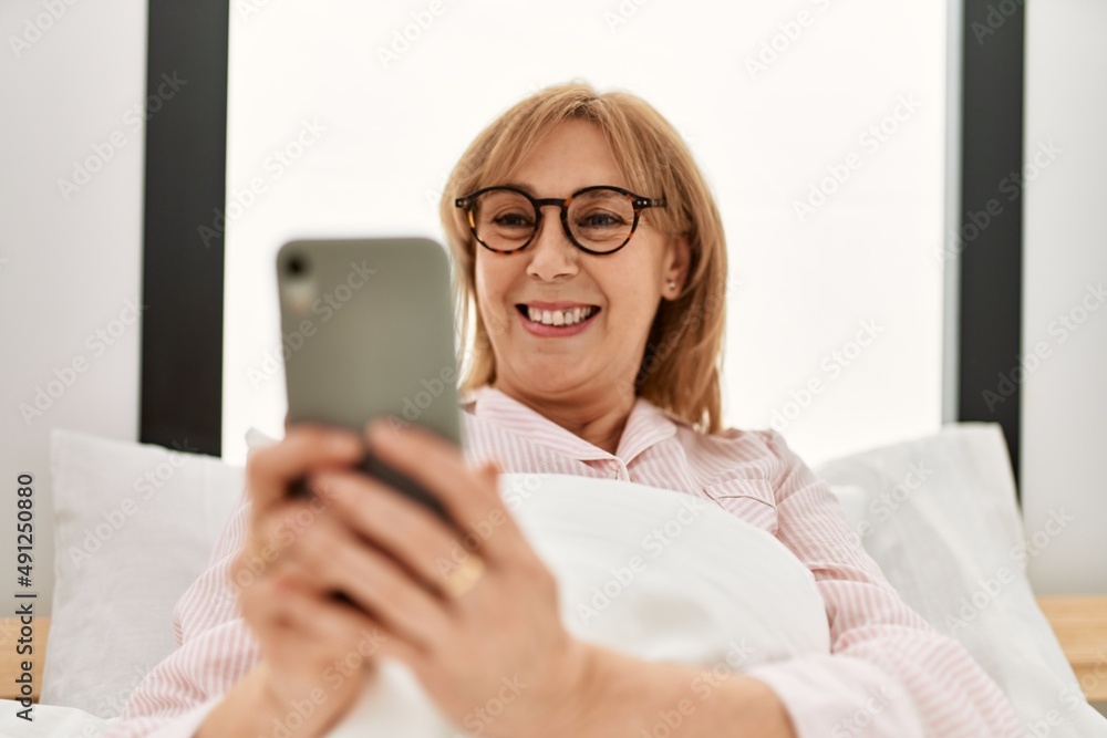 Middle age blonde woman using smartphone lying on the bed at home.