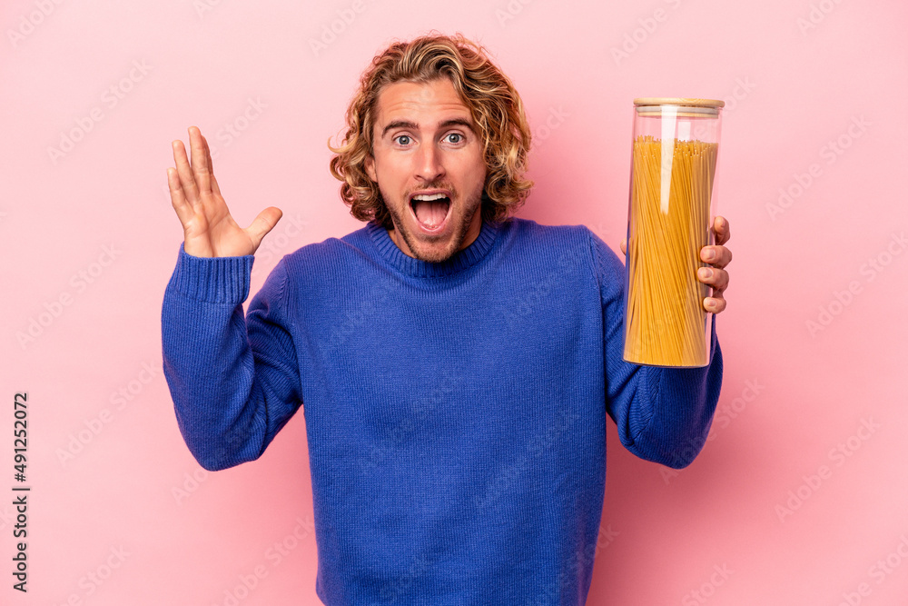 Young caucasian man holding spaghettis jar isolated on pink background receiving a pleasant surprise, excited and raising hands. Young caucasian man holding spaghettis jar isolated on pink background receiving a pleasant surprise, excited and raising hands.