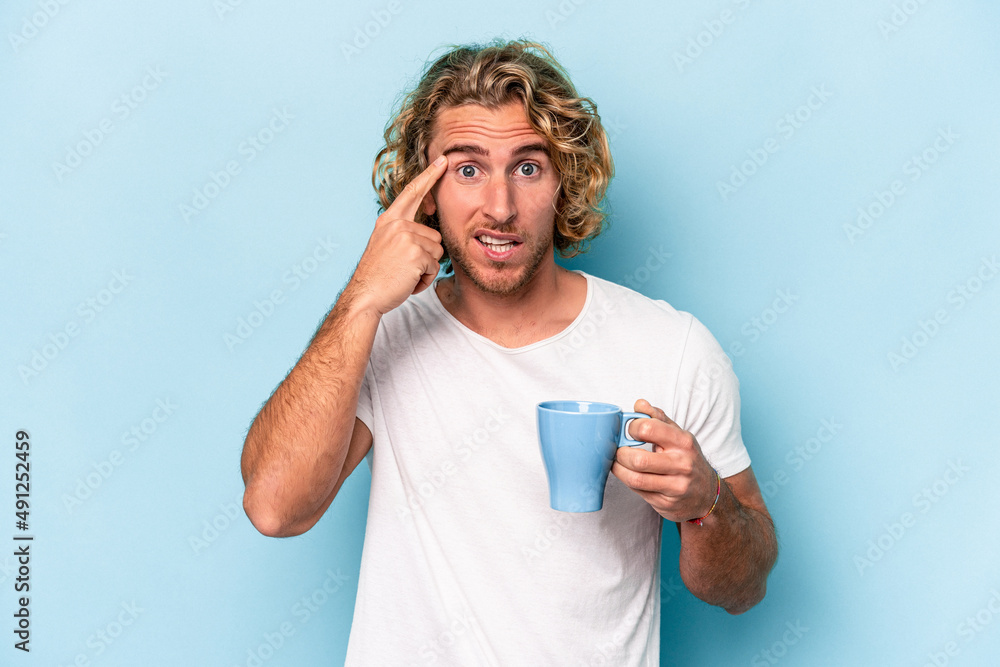 Young man holding a mug isolated on blue background showing a disappointment gesture with forefinger. Young man holding a mug isolated on blue background showing a disappointment gesture with forefinger.