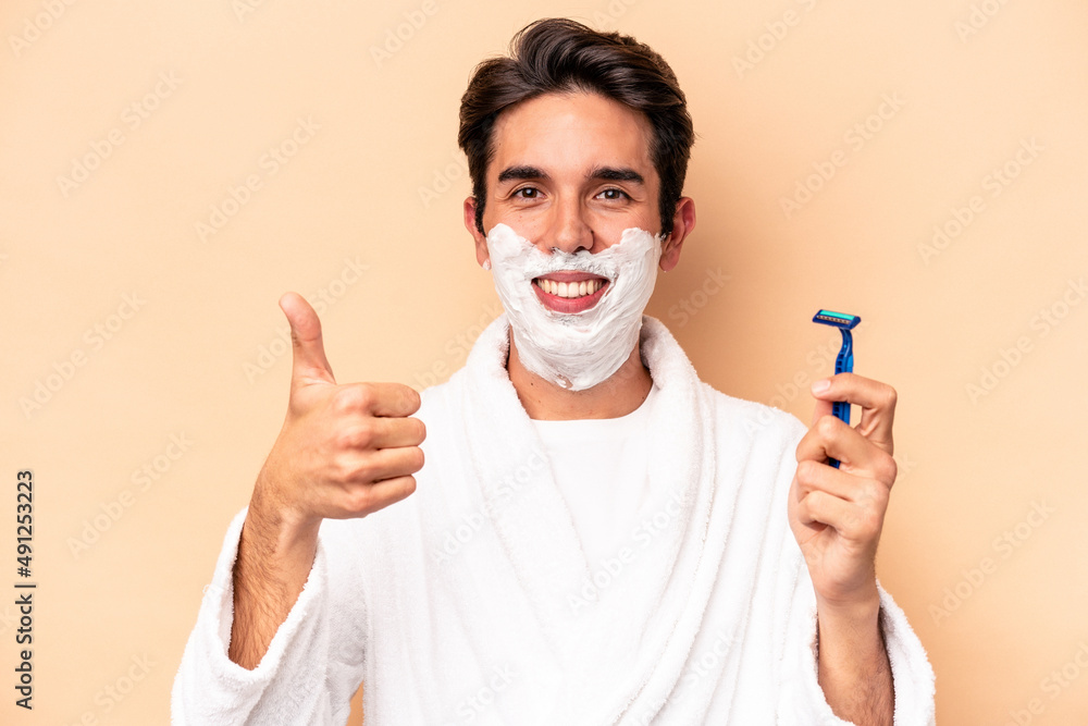 Young caucasian man shaving his beard isolated on beige background smiling and raising thumb up
