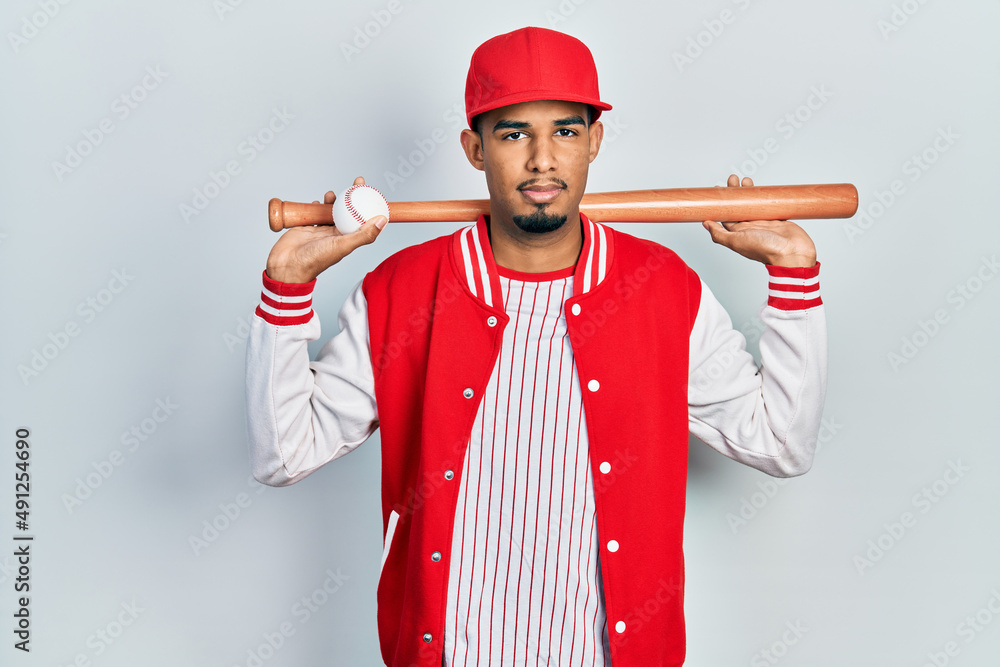 Young african american man playing baseball holding bat and ball relaxed with serious expression on face. simple and natural looking at the camera. Young african american man playing baseball holding bat and ball relaxed with serious expression on face. simple and natural looking at the camera.