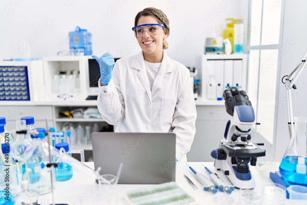 Young hispanic woman wearing scientist uniform working at laboratory pointing thumb up to the side smiling happy with open mouth Young hispanic woman wearing scientist uniform working at laboratory pointing thumb up to the side smiling happy with open mouth