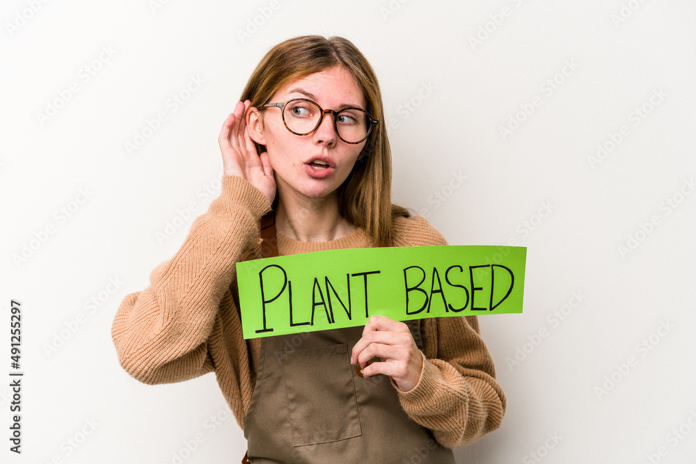 Young gardener woman holding a plan based placard isolated on white background trying to listening a gossip. Young gardener woman holding a plan based placard isolated on white background trying to listening a gossip.