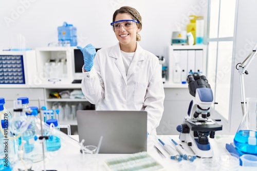 Young hispanic woman wearing scientist uniform working at laboratory pointing thumb up to the side smiling happy with open mouth