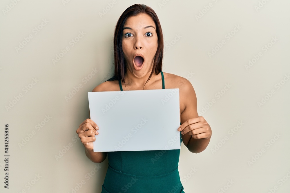 Young latin woman holding blank empty banner scared and amazed with open mouth for surprise, disbelief face Young latin woman holding blank empty banner scared and amazed with open mouth for surprise, disbelief face