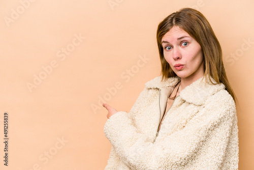 Young English woman isolated on beige background smiling and pointing aside, showing something at blank space. © Asier