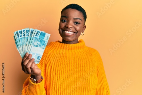 Young african american woman holding 100 brazilian real banknotes looking positive and happy standing and smiling with a confident smile showing teeth photo