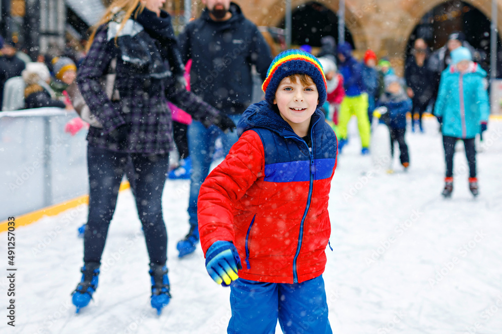 Happy little kid boy in colorful warm clothes skating on a rink of Christmas market or fair. Healthy child having fun on ice skate. Lot of people celebrating holiday and having active winter leisure Happy little kid boy in colorful warm clothes skating on a rink of Christmas market or fair. Healthy child having fun on ice skate. Lot of people celebrating holiday and having active winter leisure