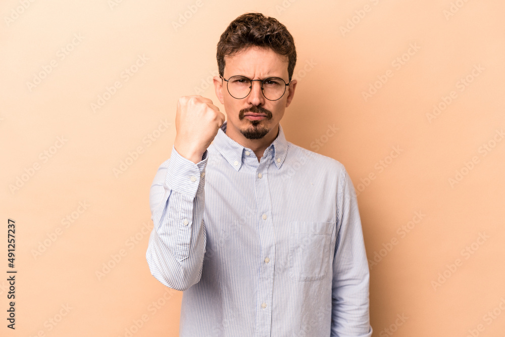 Young caucasian man isolated on beige background showing fist to camera, aggressive facial expression. Young caucasian man isolated on beige background showing fist to camera, aggressive facial expression.