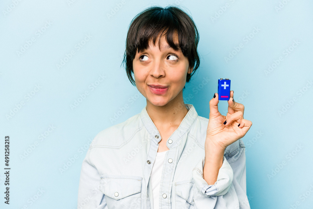 Young hispanic woman holding a batterie isolated on blue background confused, feels doubtful and unsure. Young hispanic woman holding a batterie isolated on blue background confused, feels doubtful and unsure.