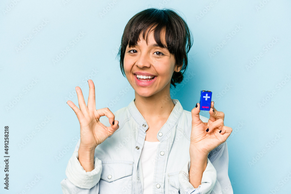 Young hispanic woman holding a batterie isolated on blue background cheerful and confident showing ok gesture. Young hispanic woman holding a batterie isolated on blue background cheerful and confident showing ok gesture.