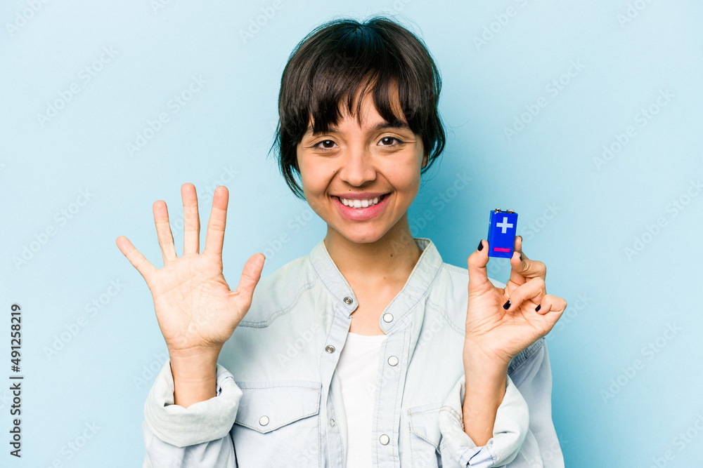 Young hispanic woman holding a batterie isolated on blue background smiling cheerful showing number five with fingers. Young hispanic woman holding a batterie isolated on blue background smiling cheerful showing number five with fingers.