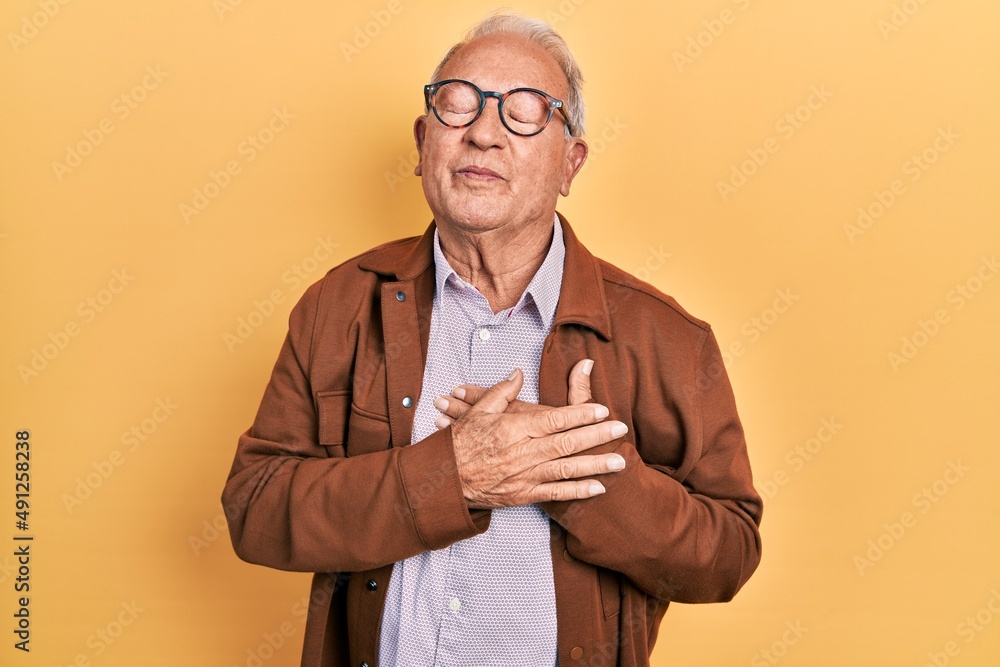 Senior man with grey hair wearing casual jacket and glasses smiling with hands on chest with closed eyes and grateful gesture on face. health concept. Senior man with grey hair wearing casual jacket and glasses smiling with hands on chest with closed eyes and grateful gesture on face. health concept.