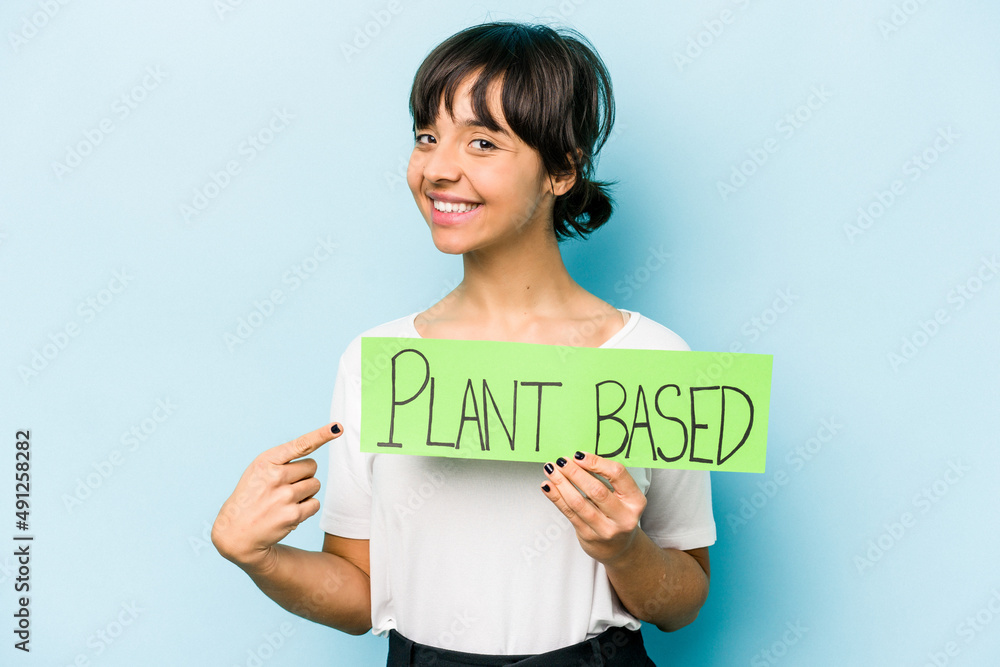 Young hispanic woman holding a plant based placard isolated on blue background smiling and pointing aside, showing something at blank space. Young hispanic woman holding a plant based placard isolated on blue background smiling and pointing aside, showing something at blank space.