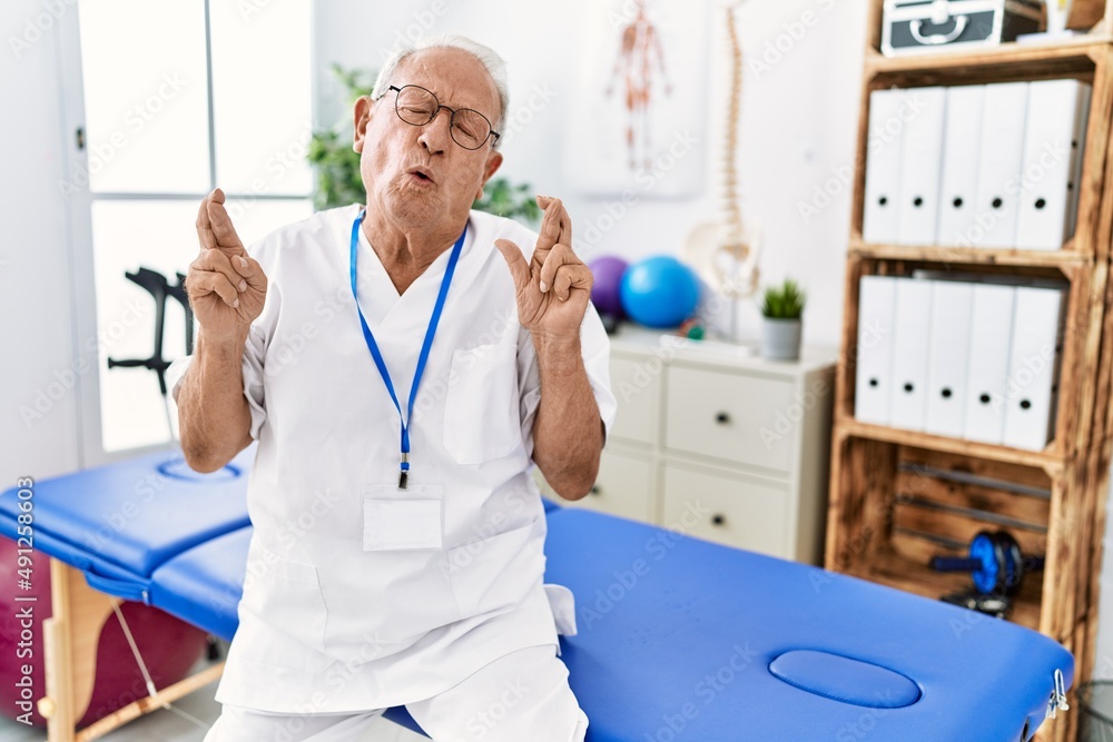 Fototapeta premium Senior physiotherapy man working at pain recovery clinic gesturing finger crossed smiling with hope and eyes closed. luck and superstitious concept.
