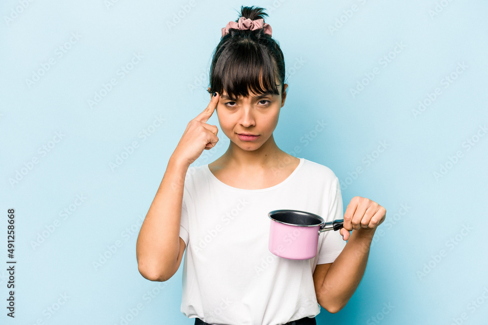 Young hispanic woman holding a saucepan isolated on blue background pointing temple with finger, thinking, focused on a task. Young hispanic woman holding a saucepan isolated on blue background pointing temple with finger, thinking, focused on a task.