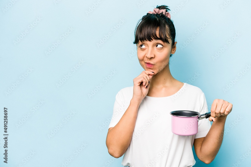 Young hispanic woman holding a saucepan isolated on blue background looking sideways with doubtful and skeptical expression. Young hispanic woman holding a saucepan isolated on blue background looking sideways with doubtful and skeptical expression.