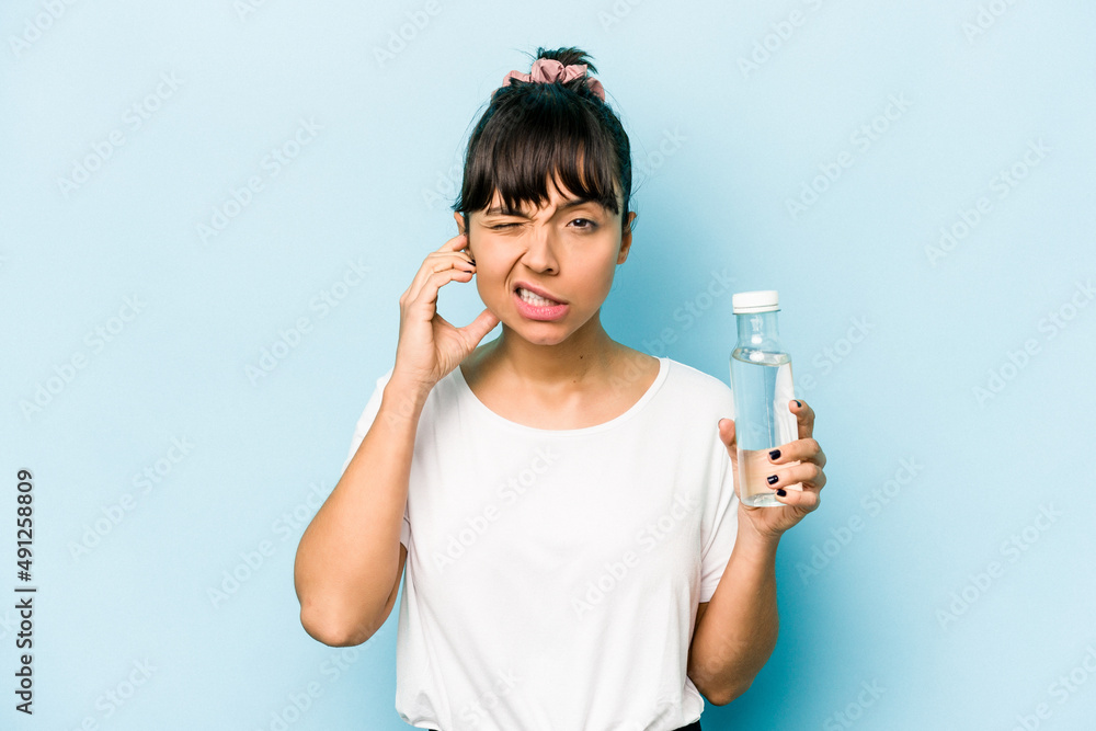 Young hispanic woman holding a bottle of water isolated on blue background covering ears with hands. Young hispanic woman holding a bottle of water isolated on blue background covering ears with hands.