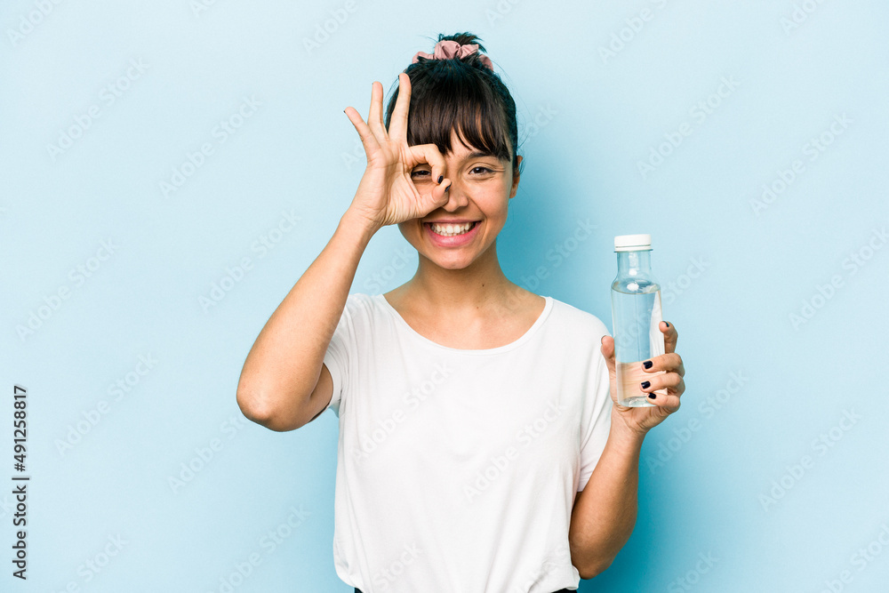 Young hispanic woman holding a bottle of water isolated on blue background excited keeping ok gesture on eye. Young hispanic woman holding a bottle of water isolated on blue background excited keeping ok gesture on eye.
