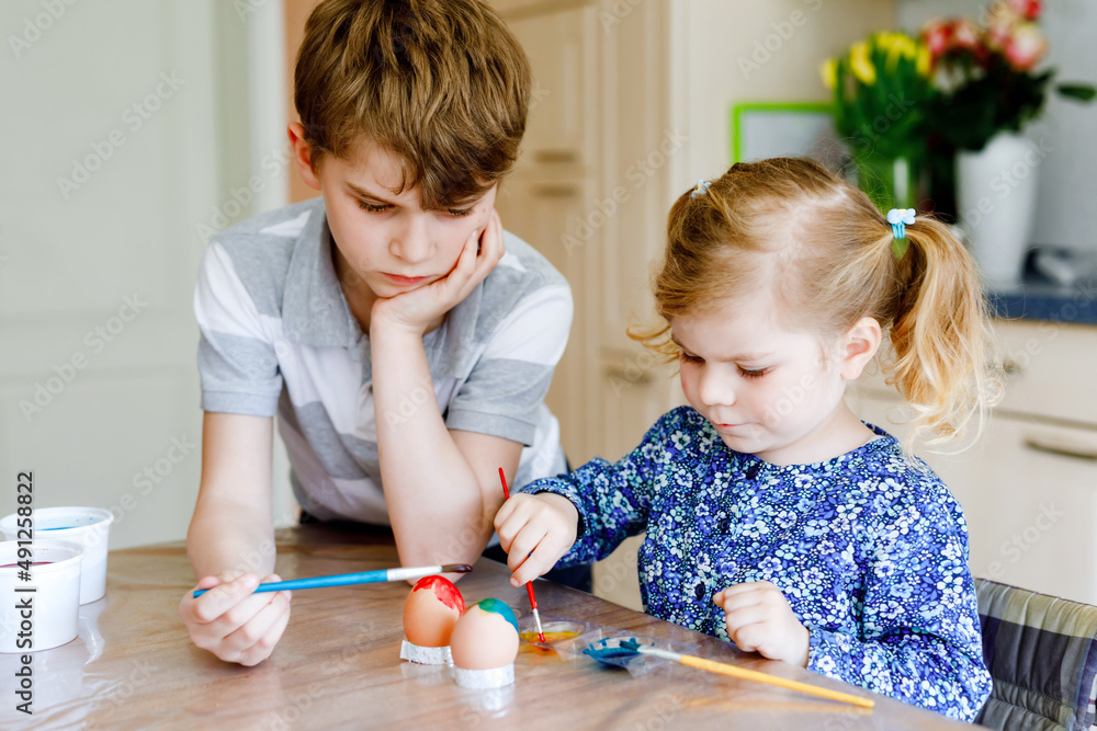 Naklejka premium Excited little toddler girl and older brother, school kid boy coloring eggs for Easter. Two cute children, siblings looking surprised at colorful eggs, celebrating holiday with family.