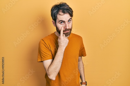 Caucasian man with beard wearing casual yellow t shirt pointing to the eye watching you gesture, suspicious expression © Krakenimages.com