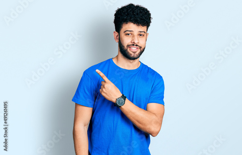 Young arab man with beard wearing casual blue t shirt pointing with hand finger to the side showing advertisement, serious and calm face © Krakenimages.com