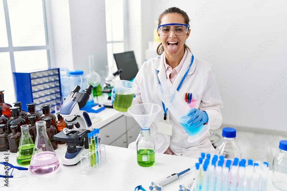 Young hispanic woman working at scientist laboratory sticking tongue out happy with funny expression. Young hispanic woman working at scientist laboratory sticking tongue out happy with funny expression.